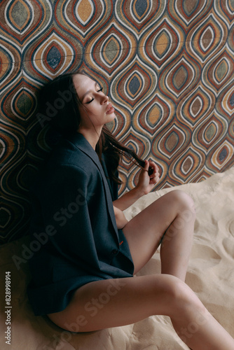 Young sexy woman in an unbuttoned jacket and bare breasts and jeans posing against the wall. Model shooting in an interior studio with sand on the floor.