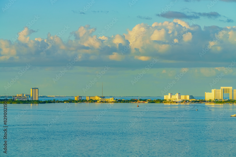 Fototapeta premium Coastal cityscape with modern buildings under a bright sky with fluffy clouds, reflecting on the calm sea. High quality photo. Nichupte Lagoon is one of the best examples of natural paradise. Sunrise