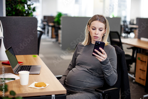 A pregnant woman sits at a desk in the office, gazing at her phone with some personal items on the table, such as a laptop and a plant, in a typical white collar worker setup