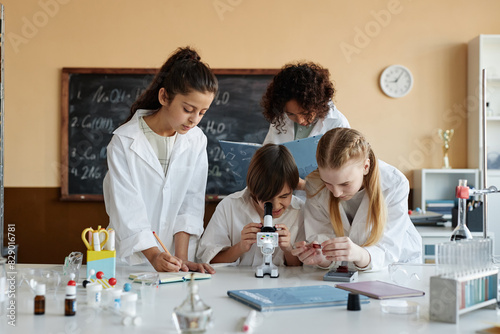 Multi-ethnic group of boys and girls wearing lab coats using microscope while doing experiment in modern Chemistry class