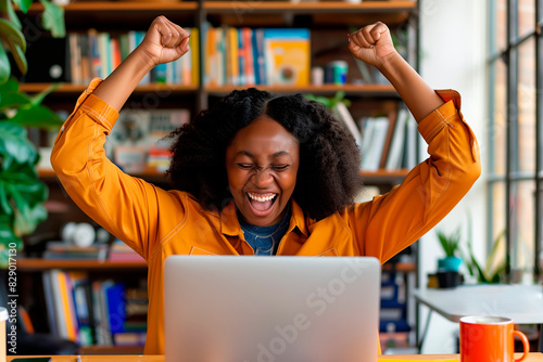 Ecstatic black woman celebrating online success with laptop in a vibrant, book-filled home office photo