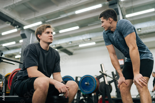 Obraz na plátně Low-angle view of of muscular personal instructor explaining technique of exercise to young beginner sportsman sitting on bench with barbell during fitness training