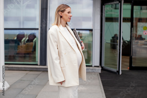 Beautiful pregnant blonde woman in a white business suit with a blazer stands outdoors with a laptop near a corporate building, showcasing professional and modern maternity style