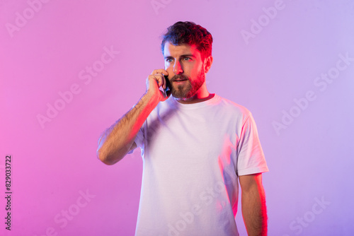 A man wearing a white t-shirt is engaged in a phone conversation, holding a cell phone to his ear. He appears focused and attentive as he communicates verbally. photo