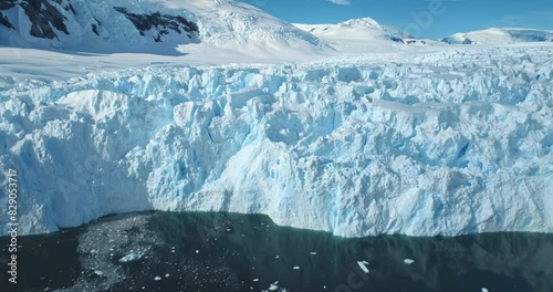 Zoom out panorama giant glacier ice wall iceberg towering on Antarctic coastline polar ocean. Sunny day blue sky, snow covered mountains. Ecology, global warming and climate change. Aerial drone shot