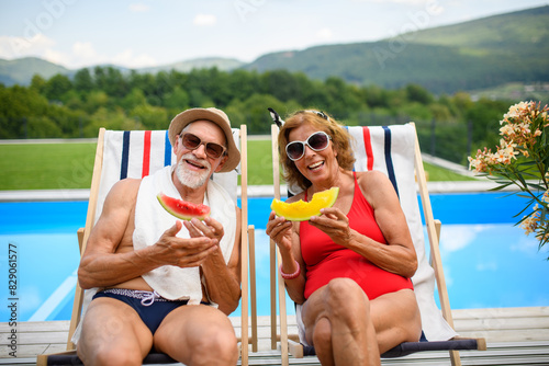 Beautiful elderly couple enjoying their vacation. Seniors having romantic time, sitting by swimming pool in lounche chairs eating watermelon. photo