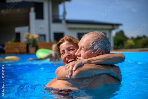 Couple of cheerful seniors shaving fun in pool with friends jumping, swiming and lounging on floats. Elderly friends spending hot day by swimming pool.