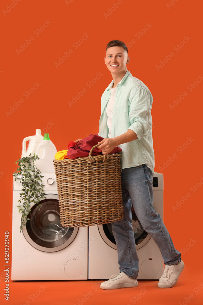 Handsome young man holding basket with laundry near washing machine on orange background Handsome young man holding basket with laundry near washing machine on orange background