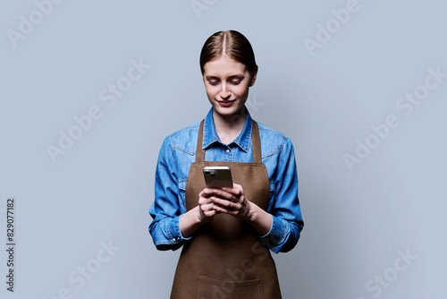 Young female worker in an apron using smartphone on gray studio background