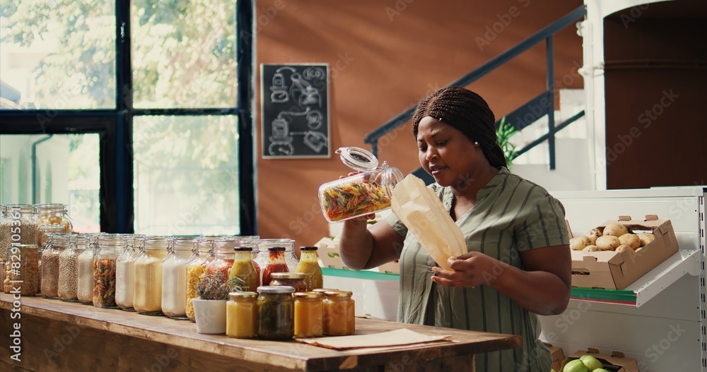 Customer filling in paper bags with bulk products, buying various pasta types and grains to restock pantry supplies. Woman enjoying grocery shopping at local farmers market. Handheld shot. Customer filling in paper bags with bulk products, buying various pasta types and grains to restock pantry supplies. Woman enjoying grocery shopping at local farmers market. Handheld shot.
