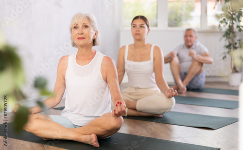 Elderly woman performs yoga exercises on mat in fitness studio with group