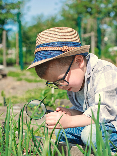 A child with a magnifying glass studies plants on the street, school assignment, biology. A boy in a hat and glasses looks at a green onion through a magnifying glass. Spring Sunny Day