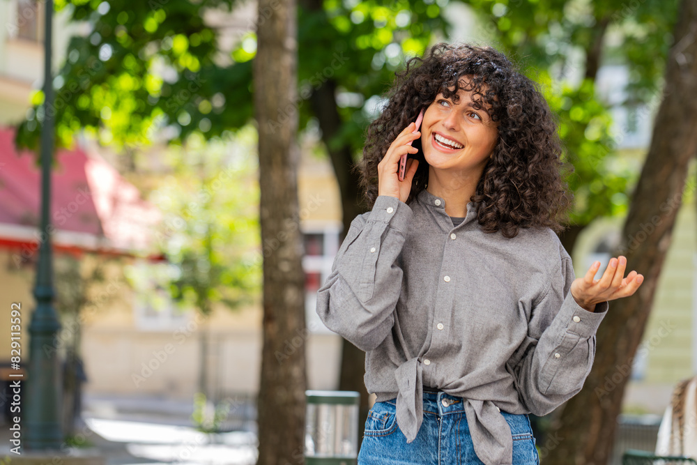 Happy Caucasian young woman having remote conversation communicate speaking by smartphone outdoors. Lady girl talking on phone unexpected good news gossip on city street. Town lifestyles. Sunny park