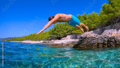LOW ANGLE VIEW: Carefree vacationer jumps head first into crystal clear seawater at Dalmatian coast. Water splashes as young man in shorts dives head first from raised rock into blue sea on summer day