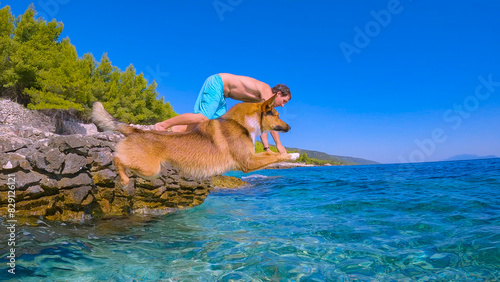 LOW ANGLE VIEW: Brown dog and young man simultaneously jump head first into refreshing crystal clear blue sea from a rocky Croatian seashore. They are enjoying summer holidays on sunny island of Hvar.