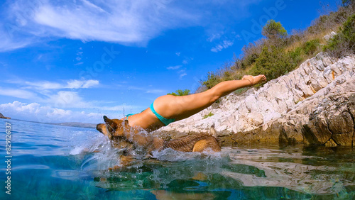 LOW ANGLE VIEW: Woman and her doggo jump off a rugged shore into crystal blue sea. Refreshing dive into blue sea with canine friend along rocky coast, embodying freedom and joy of summer adventures.