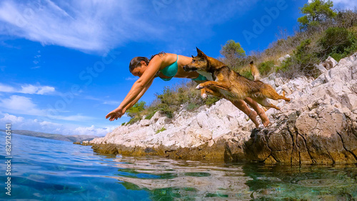 LOW ANGLE VIEW: Woman in bikini and her adorable dog in perfect synchrony as they leap from a raised rock into the sparkling blue sea, embodying freedom and the joy of summer adventures at the seaside