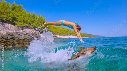 LOW ANGLE VIEW: Lady and her loyal pet enjoying a shared leap into refreshing ocean. Splashing dip into blue sea with canine friend along rocky coast, embodying freedom and joy of summer adventures.