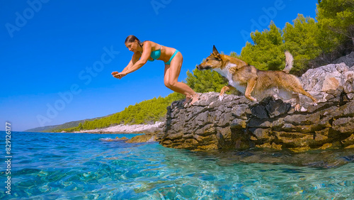 LOW ANGLE VIEW: Woman and her mixed breed dog jump off a rock into the blue sea to refresh themselves on a hot and sunny day during summer holidays. Joyful water activities on rocky coast of Hvar.