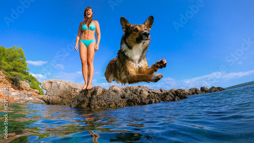 Konstfotografi LOW ANGLE VIEW: Doggo caught in the air while jumping in refreshing blue sea with a lady in bikini standing behind on a rocky shore