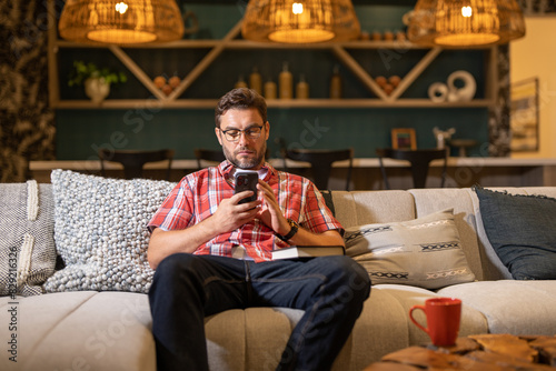 Handsome cheerful man indoors at home on sofa using mobile phone. Attractive caucasian man resting on sofa in apartment at night texting on call phone, surfing internet on smartphone. © Volodymyr