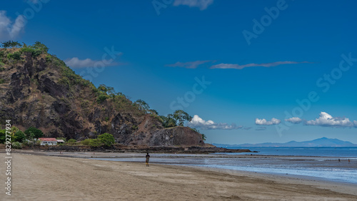 A woman walks along the ocean shore on a sandy beach at low tide. A hill against a blue sky and clouds. A white cottage is visible at the foot. Mountains on the horizon. Madagascar. Nosy Be