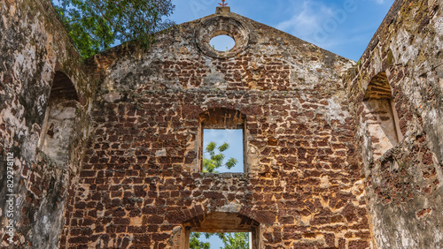 An ancient dilapidated fortress, a fragment. Weathered brick walls against a blue sky background. Tree branches are visible through the window openings. Kota a Famosa. Malaysia. Malacca.