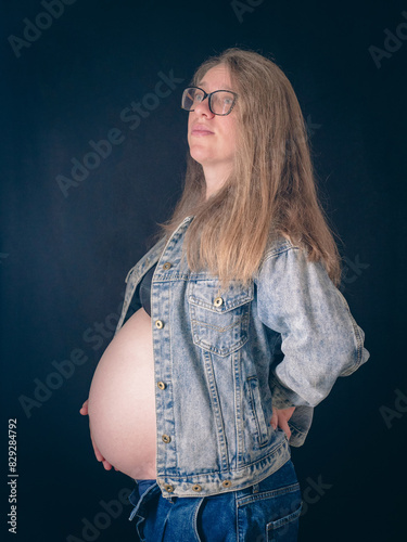 Mum-to-be wearing glasses and casual denim shows off her bump against a dark background. Long hair and serene expression emphasise the beauty of motherhood. Belt portrait in the studio.