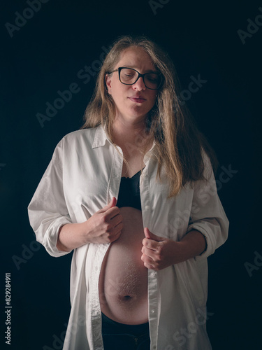 An expectant mother during her pregnancy. She is wearing a white unbuttoned shirt to show off her baby bump. The lighting emphasizes the contours of her belly.