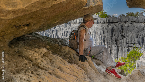 A tired man with a backpack is resting while climbing. A tourist is sitting in a crevice of rocks. Sheer karst cliffs with furrowed slopes and sharp peaks are visible through the gap. The blue sky. 