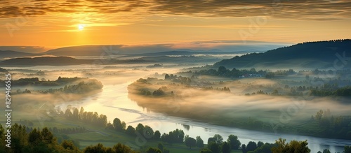 A stunning sunrise or sunset can be seen over an enchanting mist covered landscape with a scenic view of the foggy morning sky above a forest and river This captivating image captures the early summe photo