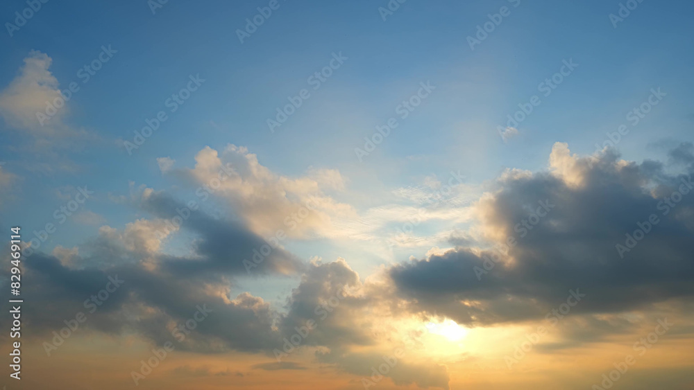 A sunset with the sun partially obscured by clouds. The sky transitions from soft blue to warm orange near the horizon, with clouds reflecting the sun's golden light. Cloud background.
