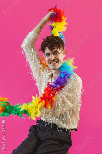 Happy non-binary person sitting with rainbow feather boa against pink background
