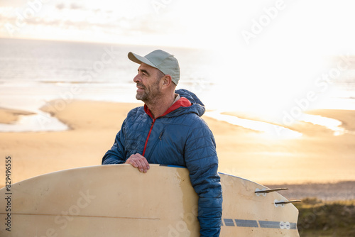 Man wearing cap and padded jacket carrying surfboard at beach photo