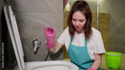 A confident brunette cleaning lady in a white T-shirt and blue apron sprays detergent from a yellow bottle on the walls of the toilet during cleaning in the bathroom in an apartment on call
