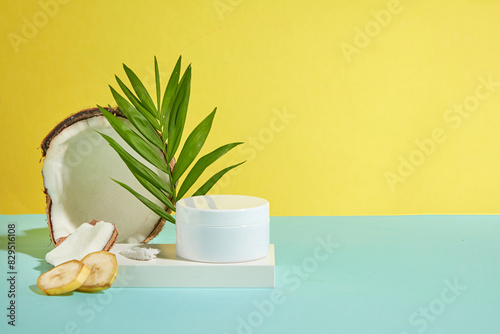 Advertising photo has fresh yellow background with tropical fruit as banana and coconut placed on the left side of green table top, a white cosmetic jar without label for adding brand packaging photo