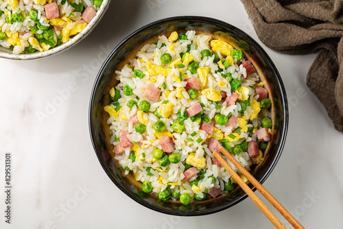 Cantonese Fried Rice with egg, green pea, ham steak, spring onion, ribe long rice in a dark bowl on a white table directly above.