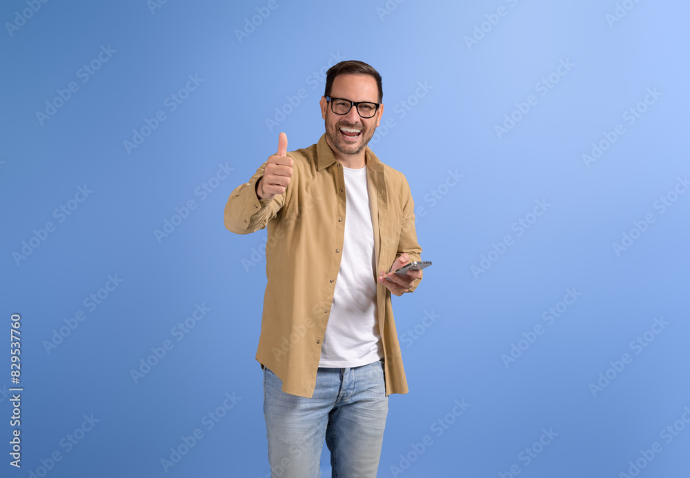 Confident happy freelancer texting over smart phone and showing thumbs up gesture on blue background Confident happy freelancer texting over smart phone and showing thumbs up gesture on blue background