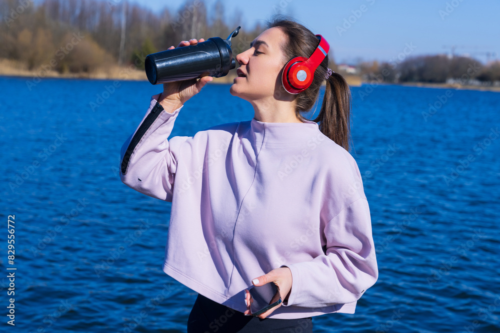 Obraz premium A young athletic woman listens to music and drinks water during breaks from training on the embankment.