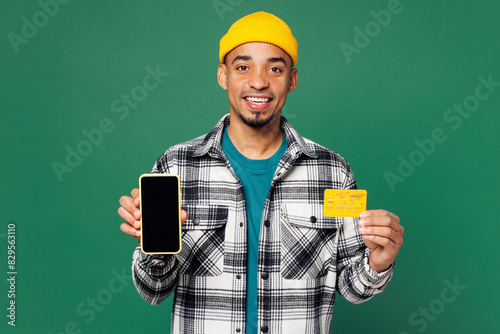 Young happy fun man wear shirt blue t-shirt yellow hat using blank screen area mobile cell phone hold credit bank card shopping online order delivery booking tour isolated on plain green background.