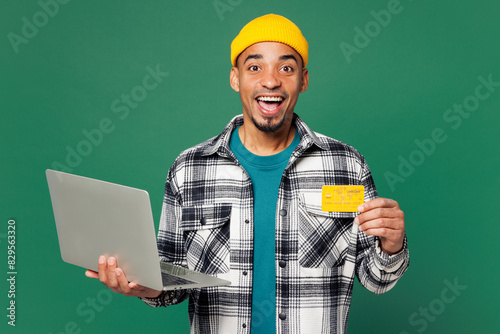 Young IT man wear shirt blue t-shirt yellow hat using laptop pc computer hold credit bank card doing online shopping order delivery booking tour isolated on plain green background. Lifestyle concept.