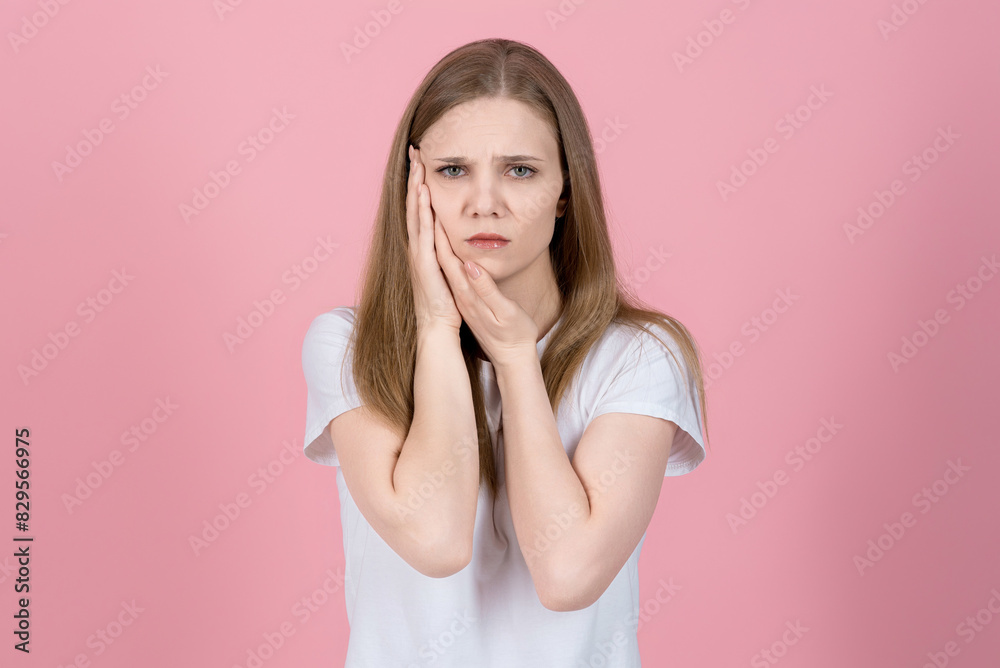 Attractive caucasian young blonde woman in casual white t-shirt looking at camera, touching cheek and suffering toothache isolated on pink studio background. Dentistry, health care