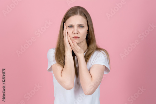 Attractive caucasian young blonde woman in casual white t-shirt looking at camera, touching cheek and suffering toothache isolated on pink studio background. Dentistry, health care