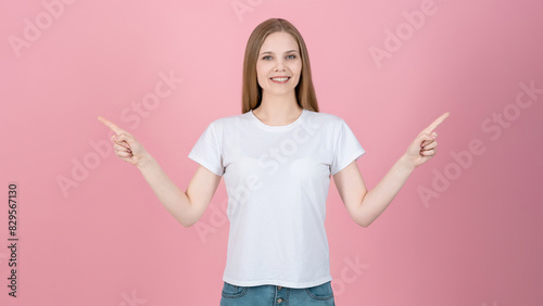 Attractive caucasian young blonde woman in casual white t-shirt smiling and pointing fingers at empty copy space for advertising on pink studio background. Sale, advice, choice.