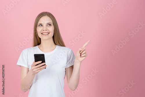 Attractive caucasian young blonde woman in casual white t-shirt using mobile phone and pointing finger at empty copy space for advertising isolated on pink studio background.