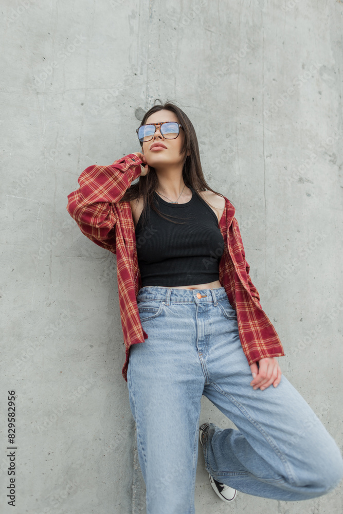 Stylish beautiful young woman model with sunglasses in a fashion shirt with jeans stands near a concrete wall on the street