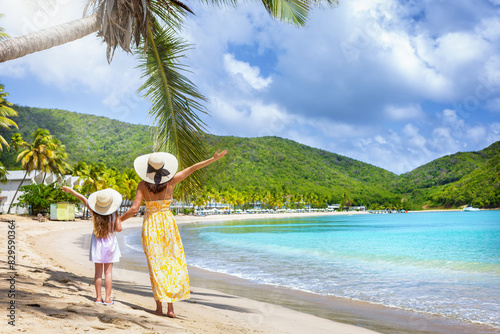 A happy mother and her daughter on summer holidays standing at a tropical beach with palm trees and turquoise sea