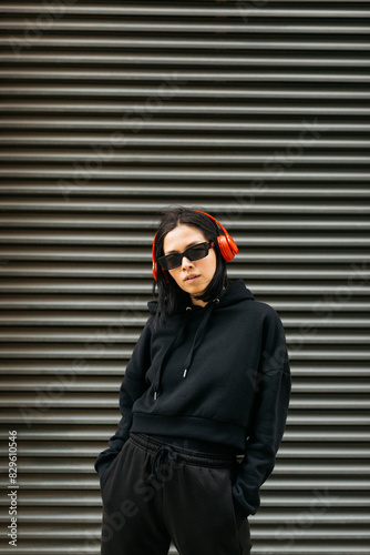 Portrait of a young woman in a black hoodie, red headphones, and sunglasses, listening music on a cool, urban street style. 