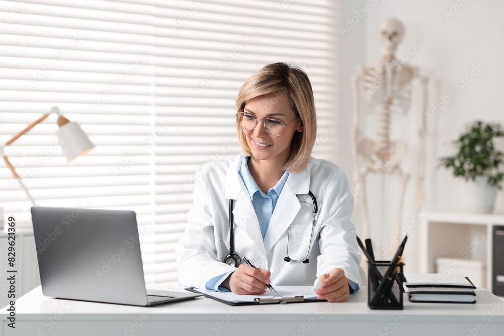 Smiling doctor with laptop having online consultation at table in office Smiling doctor with laptop having online consultation at table in office