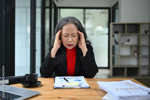 Overworked senior businesswoman massaging head feeling stress at workplace